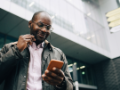 Man with glasses and earbuds looking at phone standing in front of building